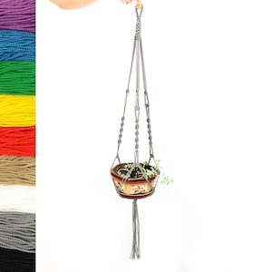 May include: Gray macrame plant hanger with a small potted plant hanging from it. The hanger is made of cotton cord and has a tassel at the bottom.