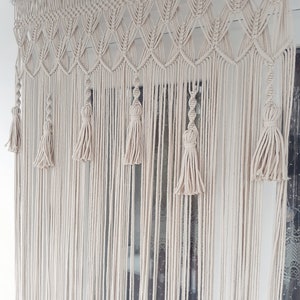 Puede incluir: Una cortina de macram&eacute; blanca con un patr&oacute;n geom&eacute;trico y borlas que cuelgan en una ventana.