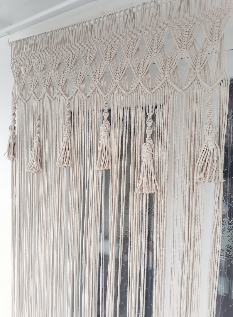 Puede incluir: Una cortina de macram&eacute; blanca con un patr&oacute;n geom&eacute;trico y borlas que cuelgan en una ventana.
