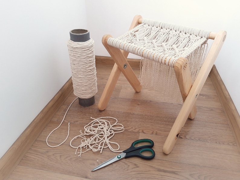 Op de afbeelding: Een houten kruk met een macram&eacute; zitting, een rol macram&eacute; koord en een schaar op een houten vloer.