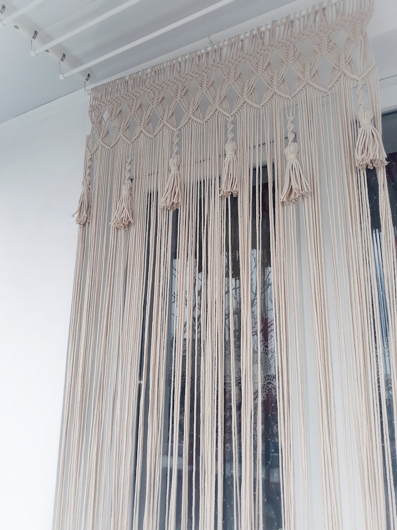 Puede incluir: Una cortina de macram&eacute; blanca con borlas cuelga de una barra de techo blanca. La cortina est&aacute; hecha de cord&oacute;n de algod&oacute;n grueso y tiene un patr&oacute;n geom&eacute;trico.