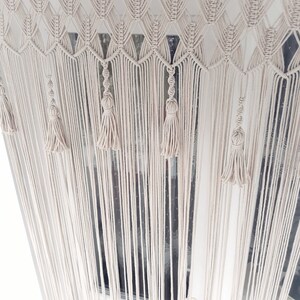Puede incluir: Una cortina de macram&eacute; blanca con un patr&oacute;n geom&eacute;trico y borlas. La cortina est&aacute; colgada en una ventana.