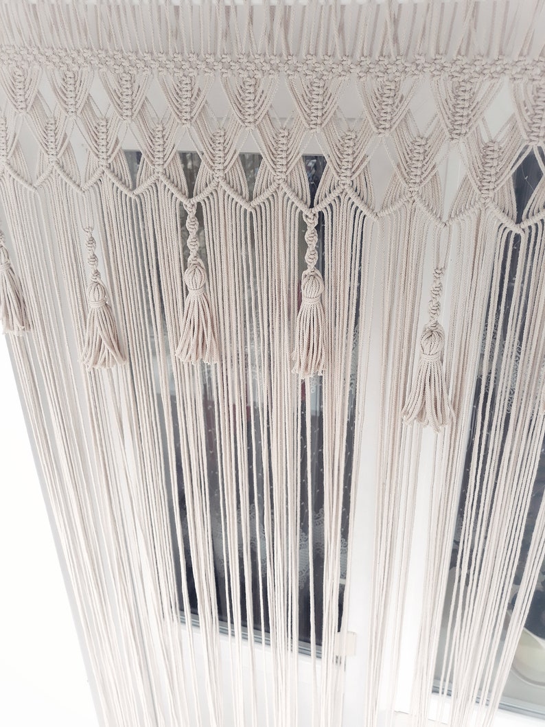 Puede incluir: Una cortina de macram&eacute; blanca con un patr&oacute;n geom&eacute;trico y borlas. La cortina est&aacute; colgada en una ventana.