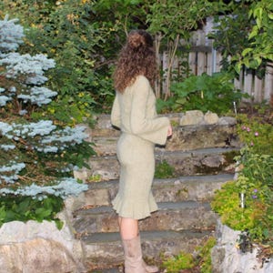 May include: A light green knit sweater and skirt set with flared sleeves and a ruffled hem. The outfit is paired with light brown boots. The person is standing on stone steps in a garden setting with lush greenery.