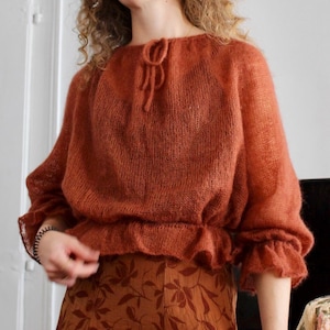 May include: A rust-colored, long-sleeved, knitted sweater with a sheer, fuzzy texture, featuring a drawstring neckline and a ruffled hem. The sweater is paired with a brown skirt that has a dark brown floral pattern printed on the fabric.