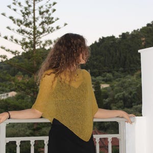 Könnte beinhalten: Ein senfgelber, locker gestrickter und halbtransparenter Poncho mit spitzem Saum, getragen über einem schwarzen Unterhemd und einem schwarzen Rock. Das Kleidungsstück hat einen weiten Ausschnitt und kurze Dolman-Ärmel aus leichtem Garn.