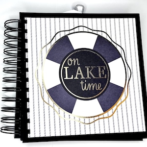 Puede incluir: Un cuaderno con espiral, tapa negra y páginas con un patrón de rayas. La portada muestra un diseño de salvavidas azul marino y blanco con las palabras "on LAKE time" en letras doradas. Un clip metálico asegura las páginas.