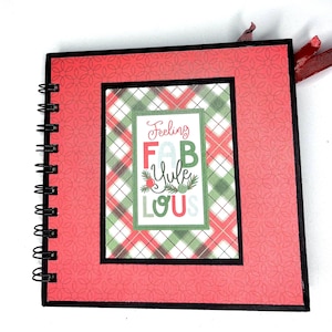 Può includere: Un album di ritagli quadrato con rilegatura a spirale e copertina rossa con un sottile motivo geometrico. Al centro presenta un inserto rettangolare con uno sfondo a quadri verdi e rossi e il testo Feeling FAB Yule LOUS.