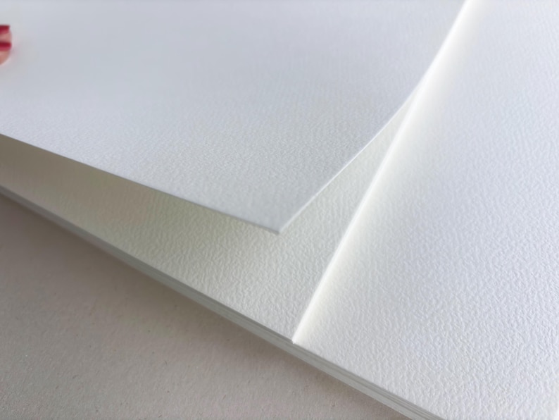 May include: Close-up of several white textured paper sheets, with a slight overlap. The paper has a subtle, grainy surface. The edges of the paper are clean and crisp. The overall impression is of a clean, minimalist aesthetic.