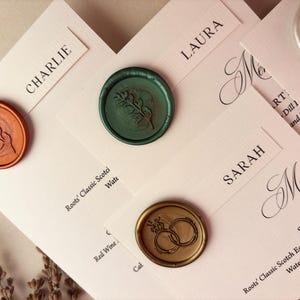 May include: Close-up of wedding stationery with name cards and menu. The cards are light pink with black text and sealed with wax stamps in shades of orange, green, and gold. The stamps feature botanical and ring designs.