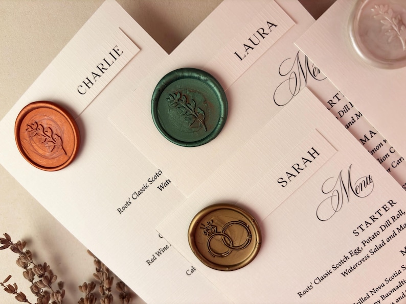 May include: Close-up of wedding stationery with name cards and menu. The cards are light pink with black text and sealed with wax stamps in shades of orange, green, and gold. The stamps feature botanical and ring designs.