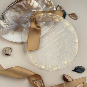 Könnte beinhalten: Eine transparente, runde Menükarte mit goldenen floralen Akzenten und Text, gebunden mit einem goldenen Band. Die Karte liegt auf einer perlmuttartigen Muschel, mit getrockneten Blättern. Das Menü enthält die Namen "LAURA & JAMIE" und das Datum "15.08.2017".