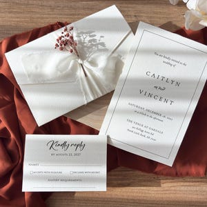 May include: A wedding invitation set comprising a cream-coloured invitation, RSVP card, and envelope. The invitation displays the names "CAITLYN and VINCENT" and event details. The envelope is tied with a white ribbon and decorated with dried flowers.