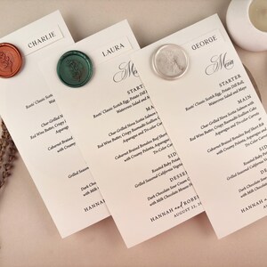 May include: Elegant wedding menu cards with names like Charlie, Laura, and George, sealed with wax stamps. The menus feature a detailed dinner selection, including starters, main courses, sides, and dessert options. The event is dated August 21, 2027.