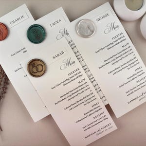 May include: Wedding menu cards with names and menu details, sealed with decorative wax stamps. The cards are white with black text, and the wax seals are in various colors, including orange, green, and silver. Two white candle holders are in the background.