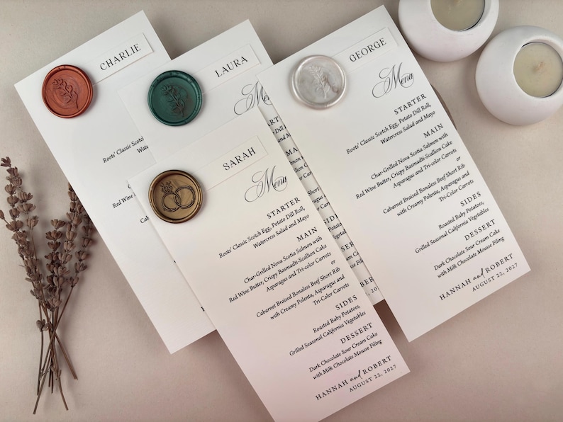 May include: Wedding menu cards with names and menu details, sealed with decorative wax stamps. The cards are white with black text, and the wax seals are in various colors, including orange, green, and silver. Two white candle holders are in the background.