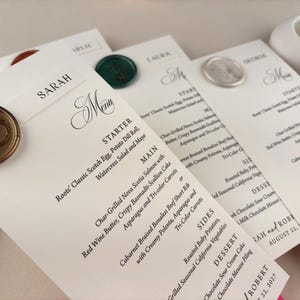 May include: Wedding menu cards with elegant calligraphy and gold wax seals. The menus feature a detailed list of dishes, including starters, main courses, sides, and desserts. The names of the guests are printed at the top of each card.