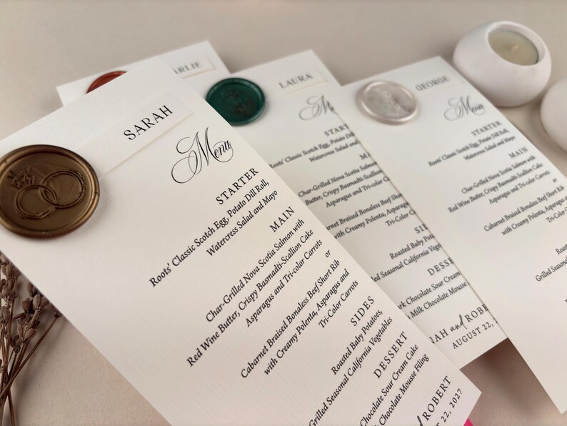 May include: Wedding menu cards with elegant calligraphy and gold wax seals. The menus feature a detailed list of dishes, including starters, main courses, sides, and desserts. The names of the guests are printed at the top of each card.