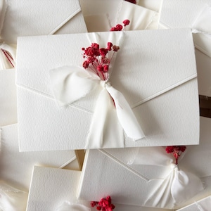 Può includere: Primo piano di buste bianche strutturate, ciascuna ornata da un nastro bianco e un grappolo di piccoli fiori rossi. Le buste sono disposte in un motivo a strati, creando un display visivamente accattivante. Tema: matrimonio o evento speciale.