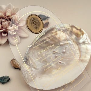 Könnte beinhalten: Eine transparente Menükarte aus Acryl mit goldenen Buchstaben und einem goldenen Wachssiegel. Die Speisekarte listet Vorspeisen, Hauptgerichte und Desserts auf. Hinter der Karte ist eine große, schillernde Muschel zu sehen, zusammen mit einer hellrosa Blume.