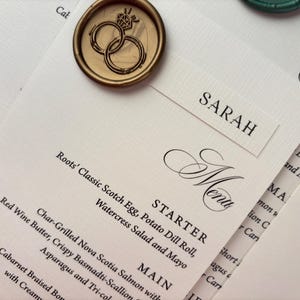 May include: Elegant wedding stationery featuring a menu with a list of dishes, a name card that says "SARAH", and a gold wax seal with intertwined rings. The menu includes the word "Menu" in a decorative font.