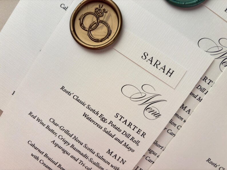 May include: Elegant wedding stationery featuring a menu with a list of dishes, a name card that says "SARAH", and a gold wax seal with intertwined rings. The menu includes the word "Menu" in a decorative font.