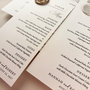 May include: Elegant wedding menu cards with detailed text, featuring starters, main courses, sides, and dessert options. The cards are adorned with decorative wax seals and the names "Hannah and Robert" with the date August 22, 2027.