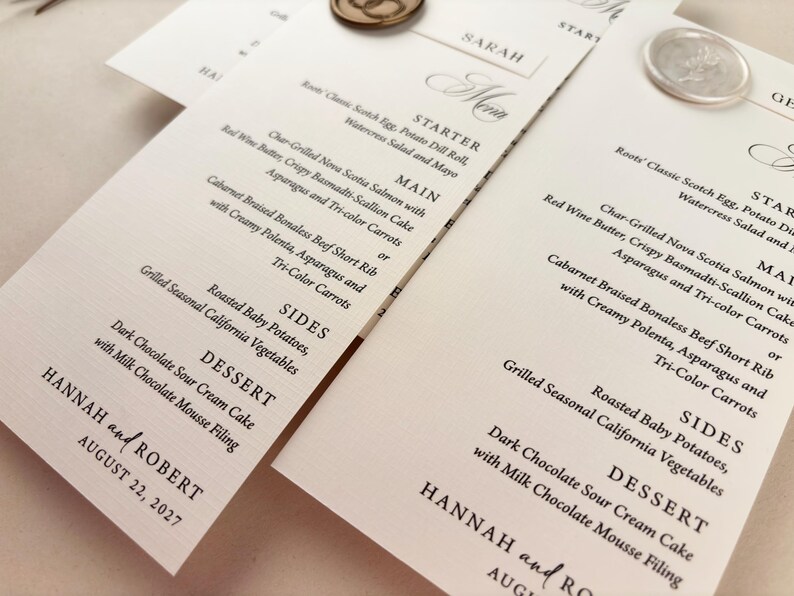 May include: Elegant wedding menu cards with detailed text, featuring starters, main courses, sides, and dessert options. The cards are adorned with decorative wax seals and the names "Hannah and Robert" with the date August 22, 2027.
