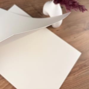 Könnte beinhalten: Ein Stapel leerer, cremefarbener Papierbögen, wobei ein Bogen teilweise angehoben ist und die Schichten freigibt. Eine kleine, weiße, abstrakte Vase mit einem Zweig getrockneter, burgunderfarbener Blumen steht im Hintergrund auf einer Holzoberfläche.