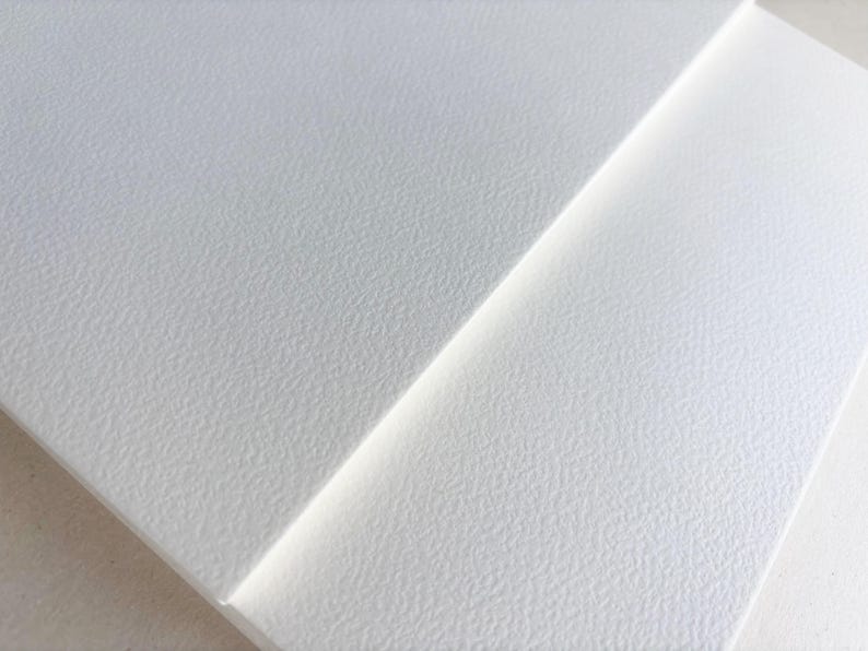 May include: Close-up of two white textured paper sheets. The sheets are angled, with one overlapping the other. The paper has a subtle, granular texture, and the lighting creates soft shadows.