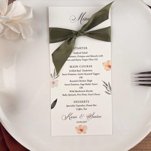 May include: A wedding menu card with a green ribbon, floral accents, and the names "Karin & Santino" with the date "16.04.2027". The menu lists starters, main courses, and desserts on a white background.