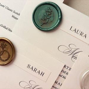 May include: Ivory place cards with names like "Laura" and "Sarah" are arranged with decorative wax seals. One seal is gold with interlocking rings, and another is teal with a sprig design. The cards are for a wedding.