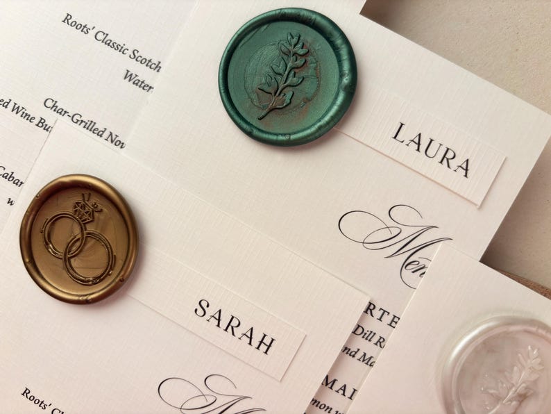 May include: Ivory place cards with names like "Laura" and "Sarah" are arranged with decorative wax seals. One seal is gold with interlocking rings, and another is teal with a sprig design. The cards are for a wedding.