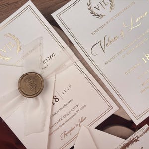 Puede incluir: Elegantes invitaciones de boda con letras y detalles dorados. Las invitaciones presentan los nombres "Valeria & Larren", la fecha "18 de octubre de 2027" y un sello de cera dorado. El diseño incluye una corona de laurel y bordes decorativos.