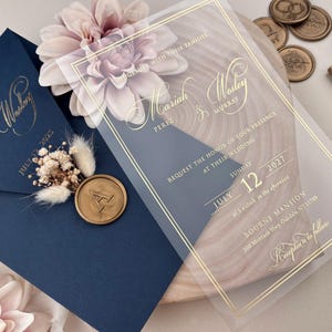 Puede incluir: Elegante juego de invitaciones de boda con un sobre azul marino con letras doradas y un sello de cera. Una invitación de acrílico transparente con letras doradas se superpone a una superficie de madera. La invitación incluye los nombres Mariah & Wesley, la fecha y el lugar.