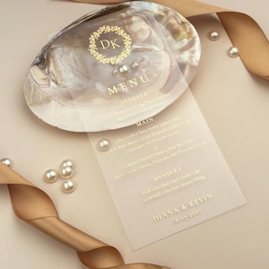 May include: A wedding menu card with gold text, featuring the names "Diana & Kevin" and the date "16.07.2017". The menu is placed on a pearlescent seashell, with pearls and a gold ribbon adding to the elegant design.