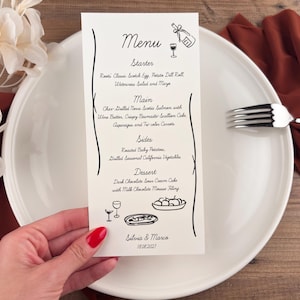 May include: A wedding menu card with a minimalist design, featuring a handwritten-style font. The menu includes starters, main courses, sides, and dessert options. A white plate, a fork, and a red napkin are also visible.