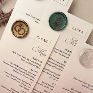 May include: Elegant wedding stationery featuring menu cards and name cards. The cards are adorned with decorative wax seals in gold, green, and silver. The menu cards list a multi-course meal.