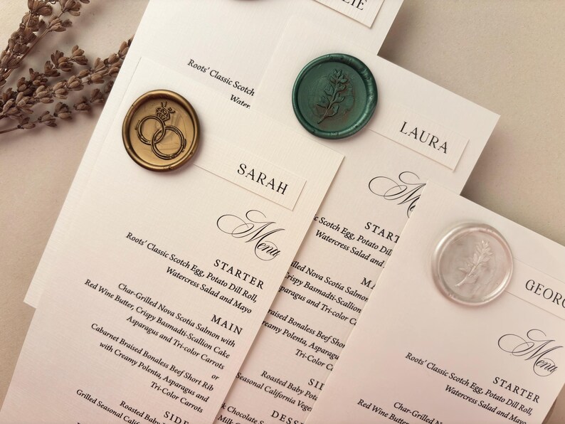 May include: Elegant wedding stationery featuring menu cards and name cards. The cards are adorned with decorative wax seals in gold, green, and silver. The menu cards list a multi-course meal.