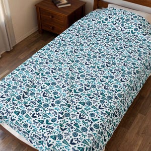 May include: A white bedspread featuring a repeating pattern of teal and navy blue graphics, including the text UNCW, pennants, pom-poms, bows, footballs, hearts, and the phrase Wings Up, covering a bed frame in a bedroom setting.