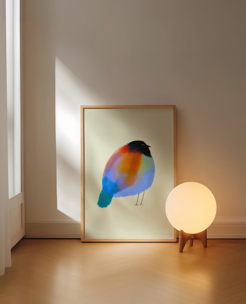 May include: A rectangular framed print featuring a stylised, round bird illustration with a black head and a vibrant, multi-coloured body in shades of orange, blue, and purple, set against a pale, neutral-toned background within a light wood frame.