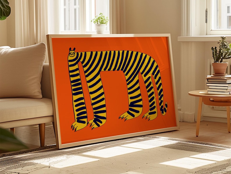 May include: Framed art print with a quirky tiger design. The artwork depicts a tiger with yellow and black stripes on an orange backdrop. The tiger has elongated legs and a simple, cartoonish style. The frame is a light wood colour.