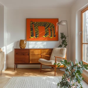 May include: A framed wall art print featuring a stylized, long-bodied creature with yellow and black stripes on a solid orange background, displayed above a wooden sideboard with a textured gold vase and a cream-colored armchair.