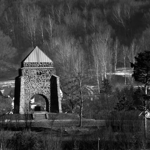May include: Black and white photograph of a stone structure with an arched entrance, resembling a tower or gatehouse. The building has a gabled roof and is surrounded by trees and a forest in the background. The image is a landscape shot.