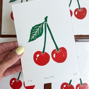 Könnte beinhalten: Eine Serie weißer Karten mit einem rot-grünen Kirschdruck. Jeder Druck zeigt zwei Kirschen mit grünem Stiel und Blatt. Das Wort "cherries" ist handschriftlich auf einer Karte. Die Drucke sind nummeriert.