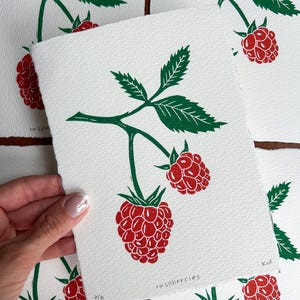 Könnte beinhalten: Ein Linolschnitt mit roten und grünen Himbeeren auf weißem Papier. Der Druck zeigt zwei reife Himbeeren mit grünen Blättern und Stielen. Das Wort "raspberries" ist in einer handschriftlichen Schriftart gedruckt. Der Druck ist auf strukturiertem Papier.