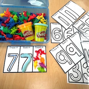 Op de afbeelding: Een blauwe plastic opbergdoos gevuld met kleurrijke miniatuur dinosaurusfiguren en twee Play-Doh-bakjes, naast verschillende witte activiteitenkaarten met cijfers en secties gelabeld Trace it, Build it en Count it, plus drie markers.