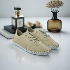 May include: A pair of beige leather sneakers with white stitching around the soles. The shoes have beige laces and are displayed with a perfume bottle and a vase of flowers.