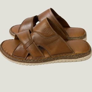 May include: A pair of brown leather sandals with a cross-strap design. The sandals have a light-coloured sole and a stitched border. The leather appears distressed, giving them a vintage look. These sandals are suitable for casual wear.