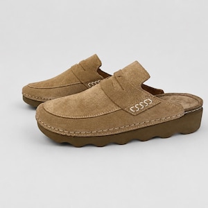 May include: A pair of tan suede slip-on loafers featuring a classic penny loafer strap across the vamp, visible light-coloured contrast stitching along the sides, and a thick, dark brown rubber sole with a distinct wavy, scalloped edge design.